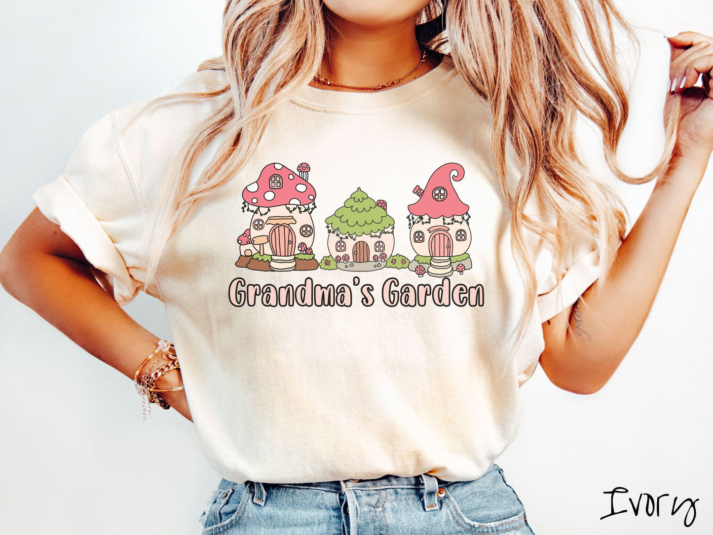 A woman wearing a cute, vintage ivory colored Comfort Colors t-shirt with the text Grandmas Garden in light pink font. Above the text are cute little cottages for gnomes made from light and dark pink mushrooms.