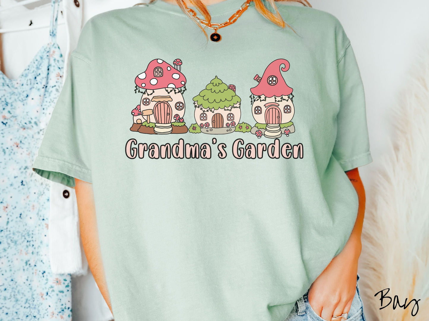 A woman wearing a cute, vintage bay colored Comfort Colors t-shirt with the text Grandmas Garden in light pink font. Above the text are cute little cottages for gnomes made from light and dark pink mushrooms.