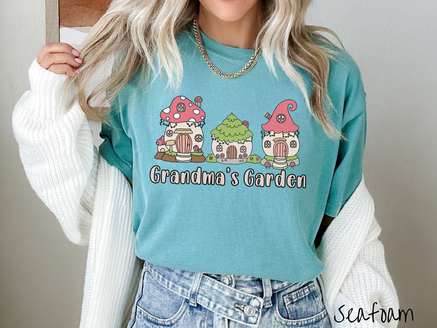 A woman wearing a cute, vintage seafoam colored Comfort Colors t-shirt with the text Grandmas Garden in light pink font. Above the text are cute little cottages for gnomes made from light and dark pink mushrooms.