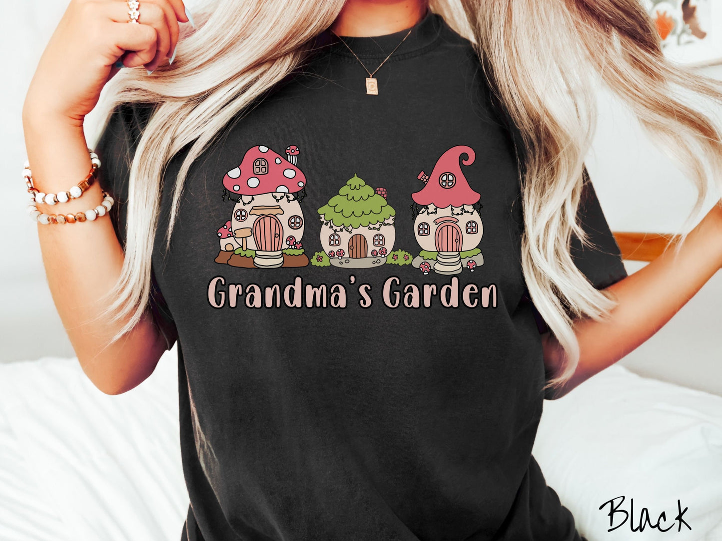 A woman wearing a cute, vintage black colored Comfort Colors t-shirt with the text Grandmas Garden in light pink font. Above the text are cute little cottages for gnomes made from light and dark pink mushrooms.