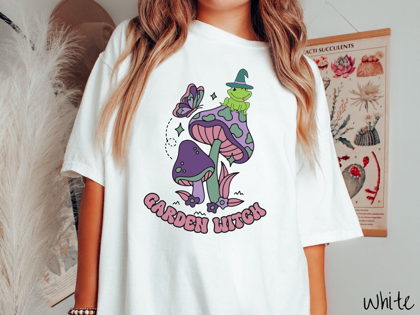 A woman wearing a cute, vintage white colored Comfort Colors t-shirt with the text Garden Witch in purple font. Above this text are two purple and green mushrooms with a green frog sitting atop one wearing a witch hat.