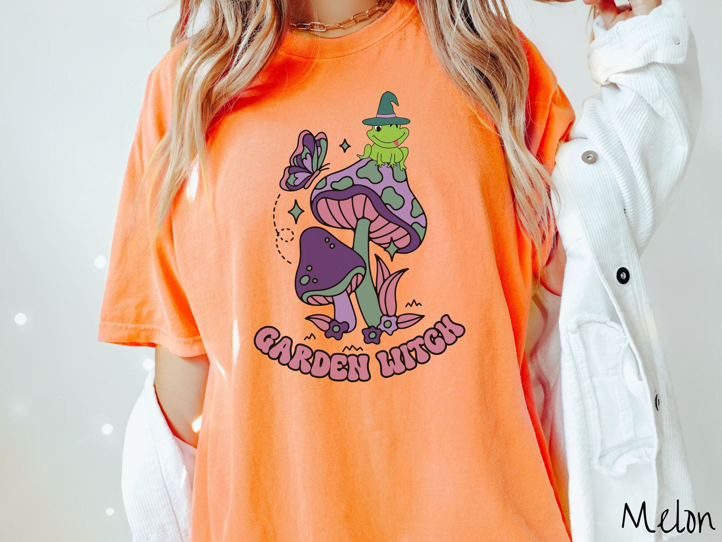 A woman wearing a cute, vintage melon colored Comfort Colors t-shirt with the text Garden Witch in purple font. Above this text are two purple and green mushrooms with a green frog sitting atop one wearing a witch hat.