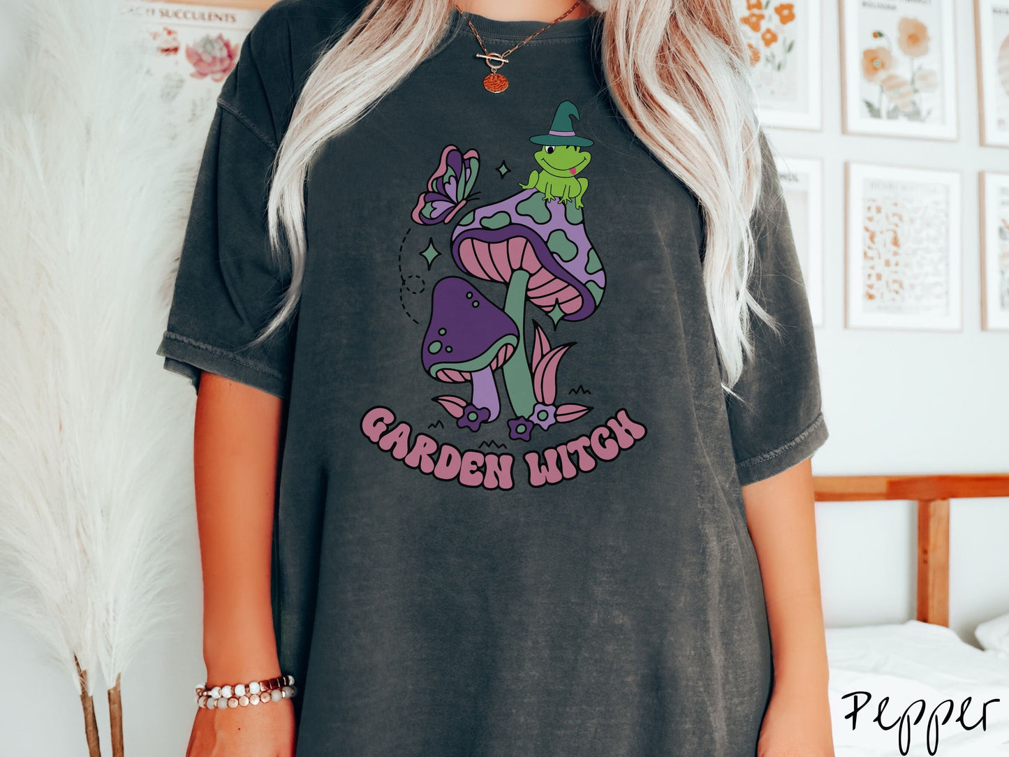 A woman wearing a cute, vintage pepper colored Comfort Colors t-shirt with the text Garden Witch in purple font. Above this text are two purple and green mushrooms with a green frog sitting atop one wearing a witch hat.