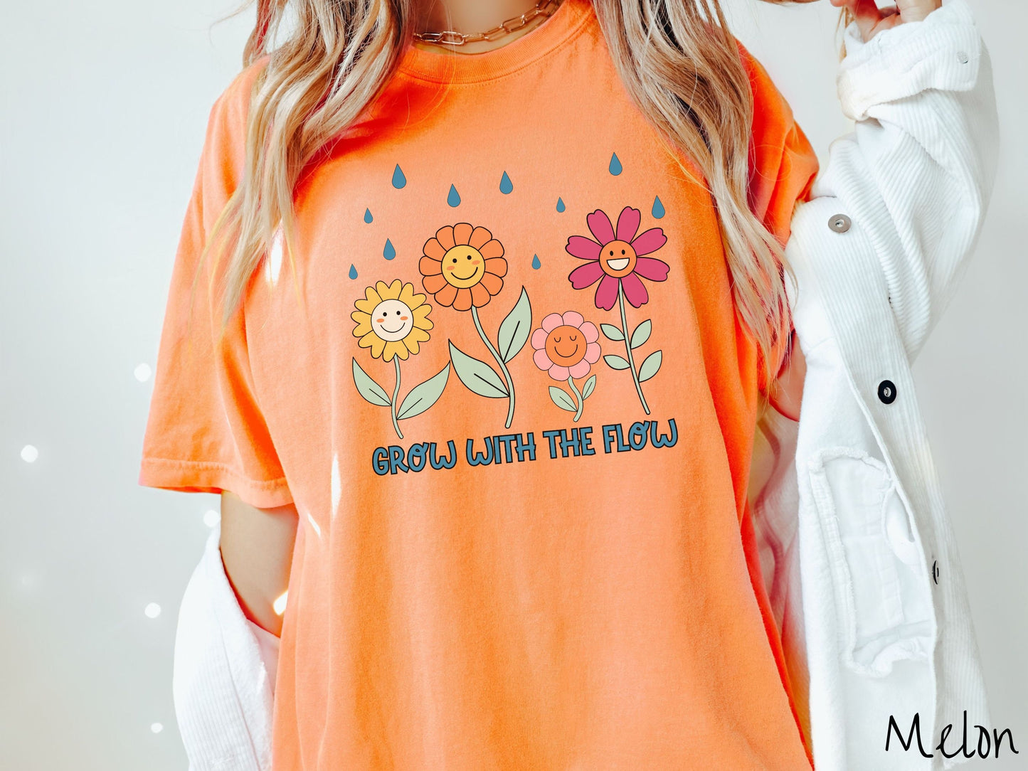 A woman wearing a cute, vintage melon colored Comfort Colors t-shirt with the text Grow with the Flow in blue font across the bottom. Above the text are smiling yellow, orange, pink, and red flowers with raindrops falling in the background.