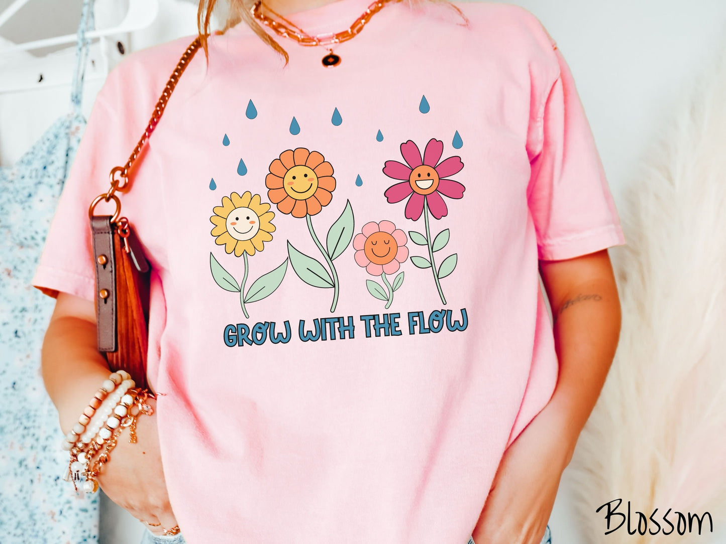 A woman wearing a cute, vintage blossom colored Comfort Colors t-shirt with the text Grow with the Flow in blue font across the bottom. Above the text are smiling yellow, orange, pink, and red flowers with raindrops falling in the background.