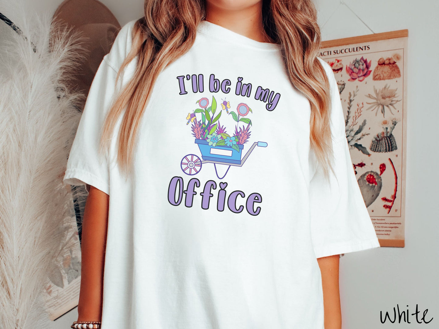 A woman wearing a cute, vintage white colored Comfort Colors t-shirt with the text I’ll Be in My Office in purple font across the front. In between the text is a blue wheelbarrow with a pink wheel stacked full of colorful plants and flowers.