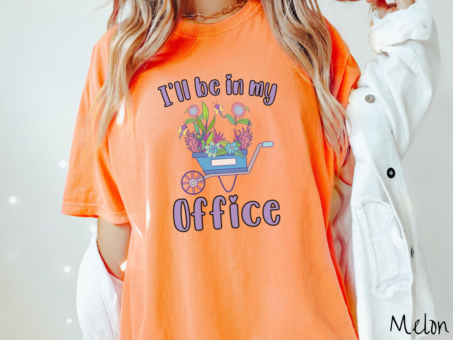 A woman wearing a cute, vintage melon colored Comfort Colors t-shirt with the text I’ll Be in My Office in purple font across the front. In between the text is a blue wheelbarrow with a pink wheel stacked full of colorful plants and flowers.