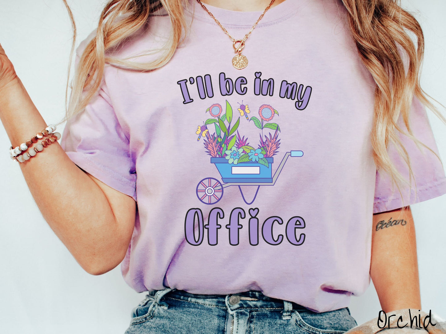 A woman wearing a cute, vintage orchid colored Comfort Colors t-shirt with the text I’ll Be in My Office in purple font across the front. In between the text is a blue wheelbarrow with a pink wheel stacked full of colorful plants and flowers.