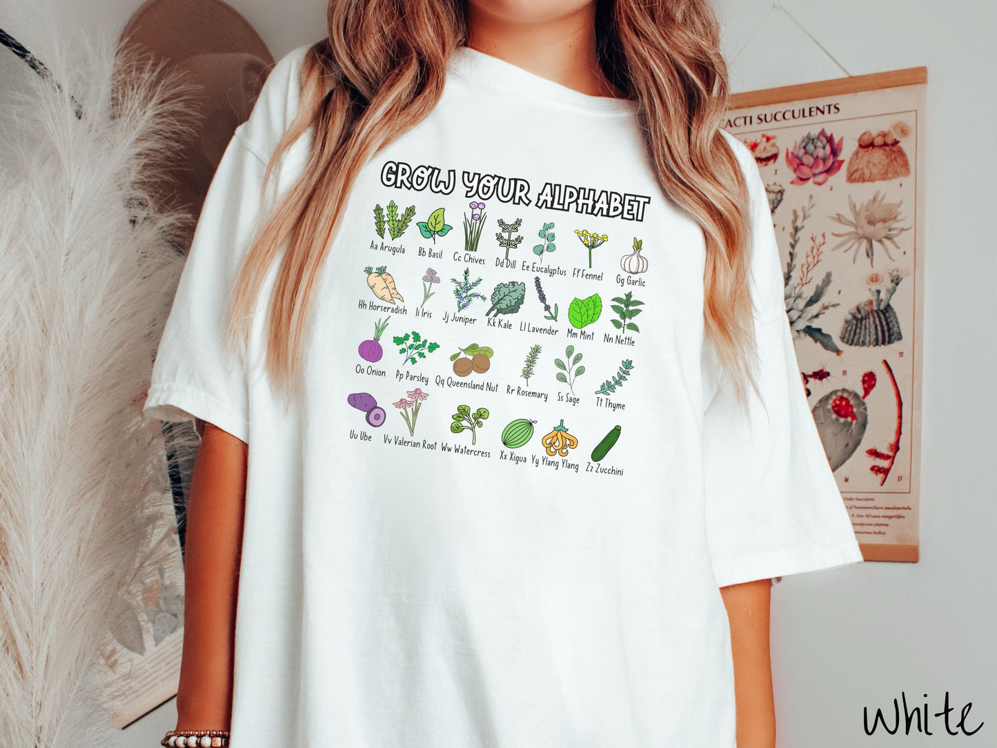 A woman wearing a cute, vintage white colored Comfort Colors t-shirt with the text Grow Your Alphabet across the top. Below this is a 4 x 7 grid of different kinds of plants and their names in alphabetical order like Arugula, Basil, and Chives.