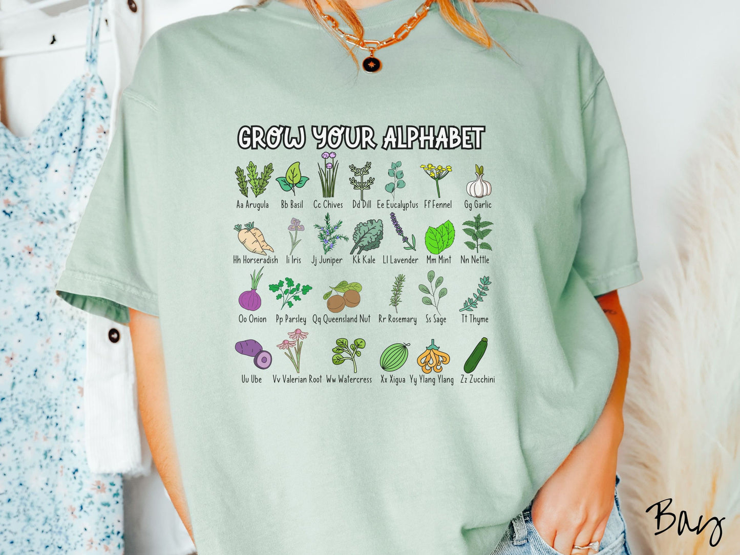 A woman wearing a cute, vintage bay colored Comfort Colors t-shirt with the text Grow Your Alphabet across the top. Below this is a 4 x 7 grid of different kinds of plants and their names in alphabetical order like Arugula, Basil, and Chives.