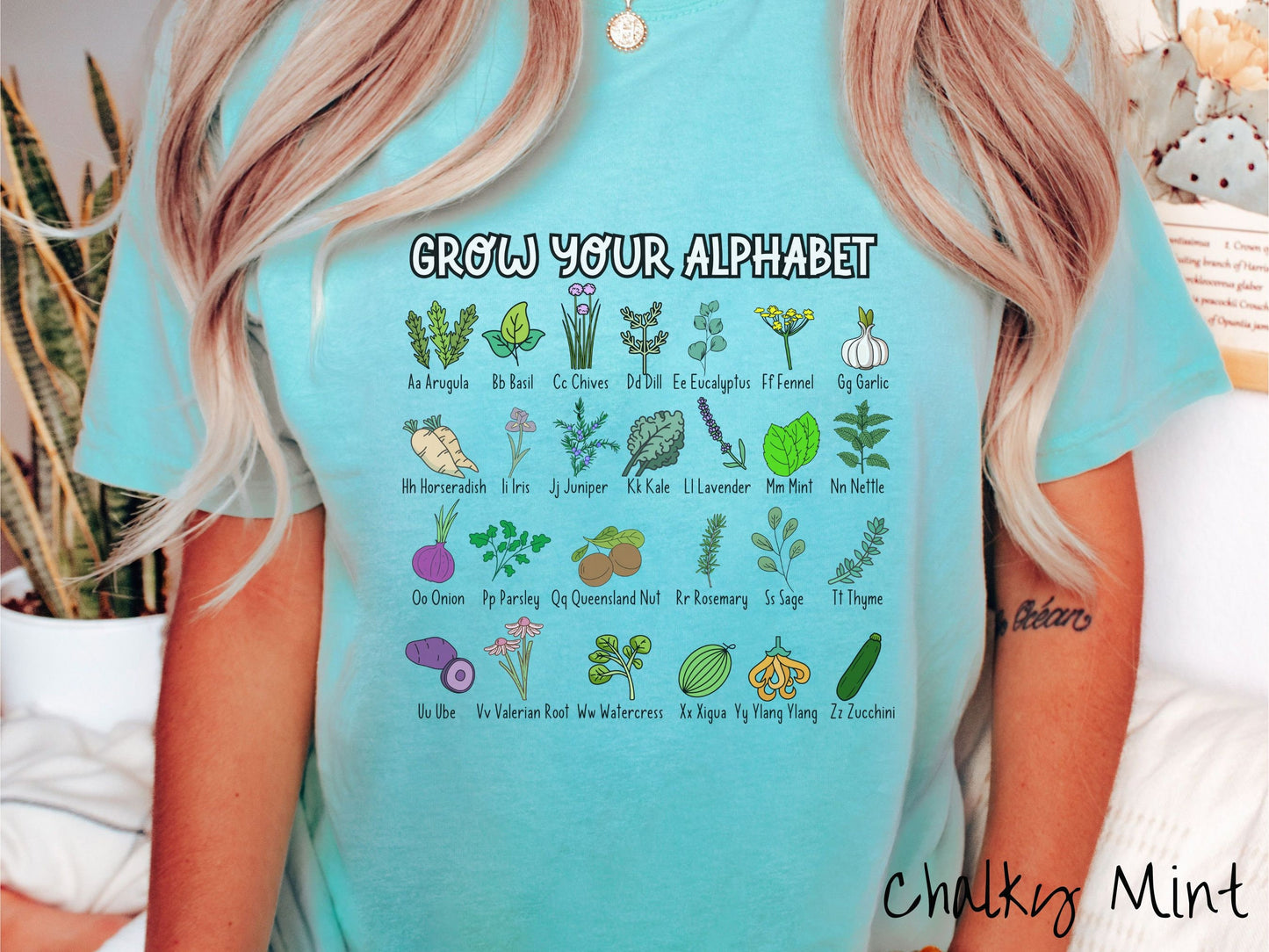 A woman wearing a cute, vintage chalky mint colored Comfort Colors t-shirt with text Grow Your Alphabet across the top. Below this is a 4 x 7 grid of different kinds of plants and their names in alphabetical order like Arugula, Basil, and Chives.
