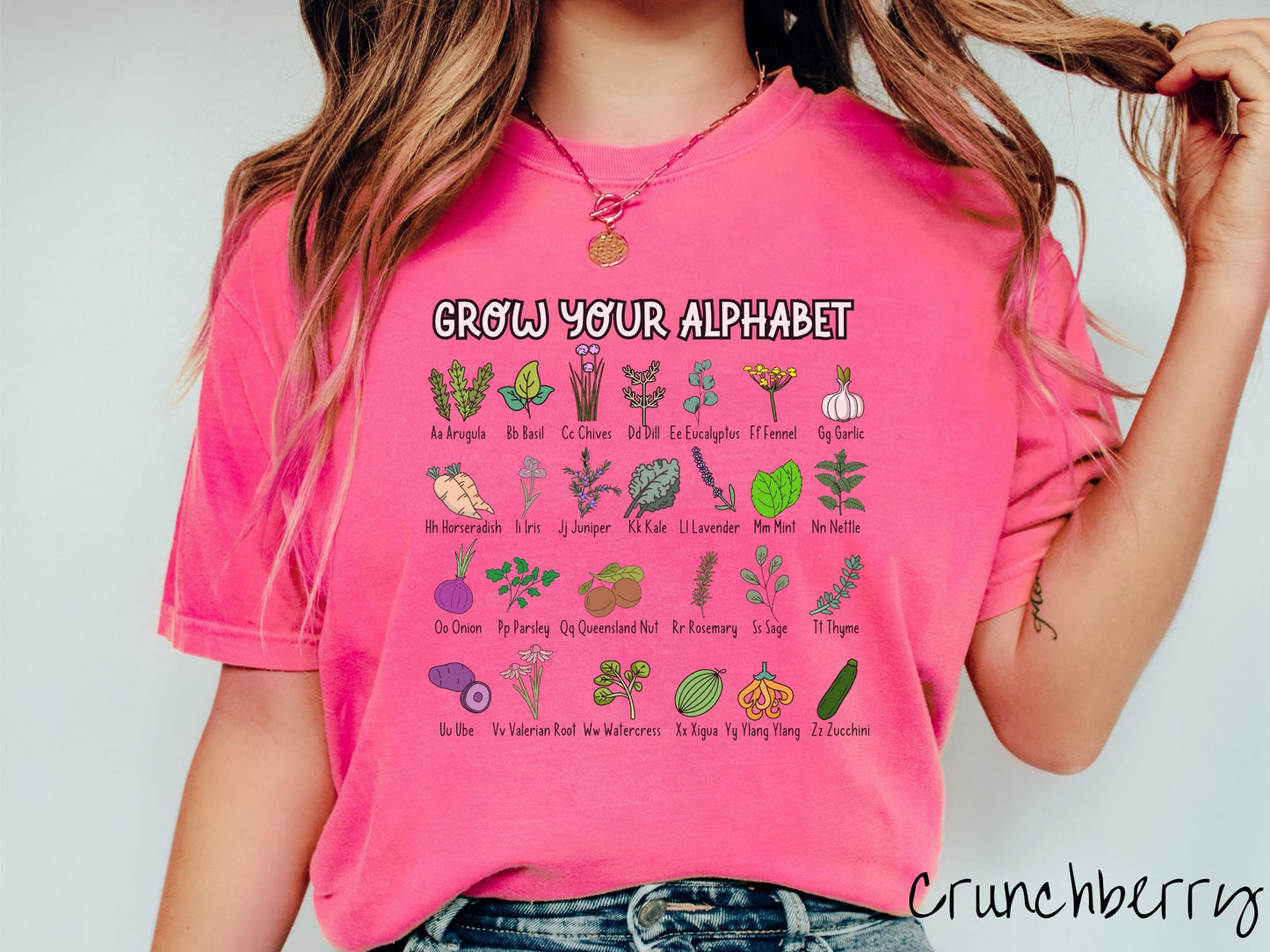 A woman wearing a cute, vintage crunchberry colored Comfort Colors t-shirt with text Grow Your Alphabet across the top. Below this is a 4 x 7 grid of different kinds of plants and their names in alphabetical order like Arugula, Basil, and Chives.