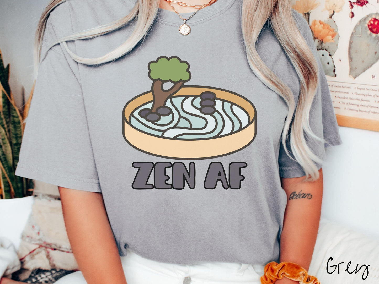 A woman wearing a cute, vintage grey colored Comfort Colors t-shirt with the text Zen AF in gray font across the font. Above this is a zen garden in a yellow container with gray and light green sand, some black pebbles, and a green tree.