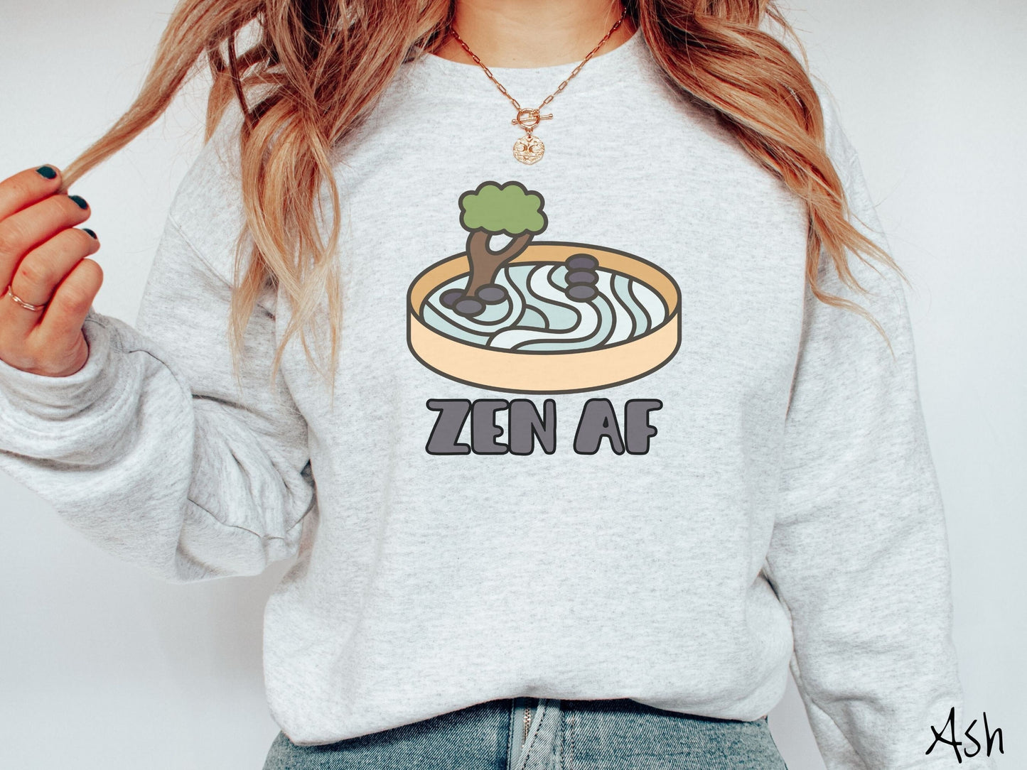 A woman wearing a cute, vintage ash colored comfy sweatshirt with the text Zen AF in gray font across the font. Above this is a zen garden in a yellow container with gray and light green sand, some black pebbles, and a green tree.