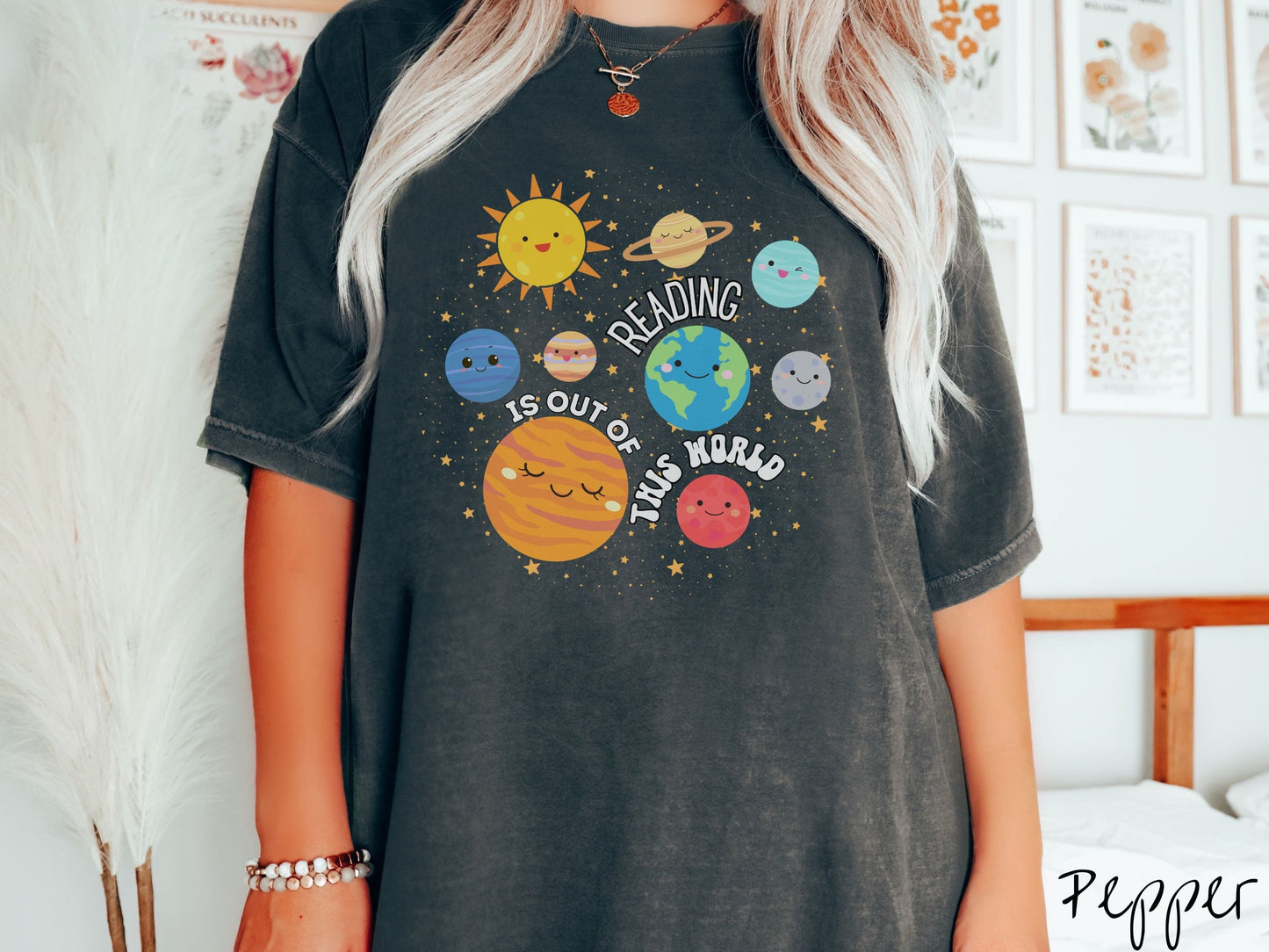 A woman wearing a cute, vintage pepper colored shirt showing the solar system and all the planets have smiling, happy faces. There is text saying Reading Is Out of This World among the planets, and there are many stars twinkling in the background.
