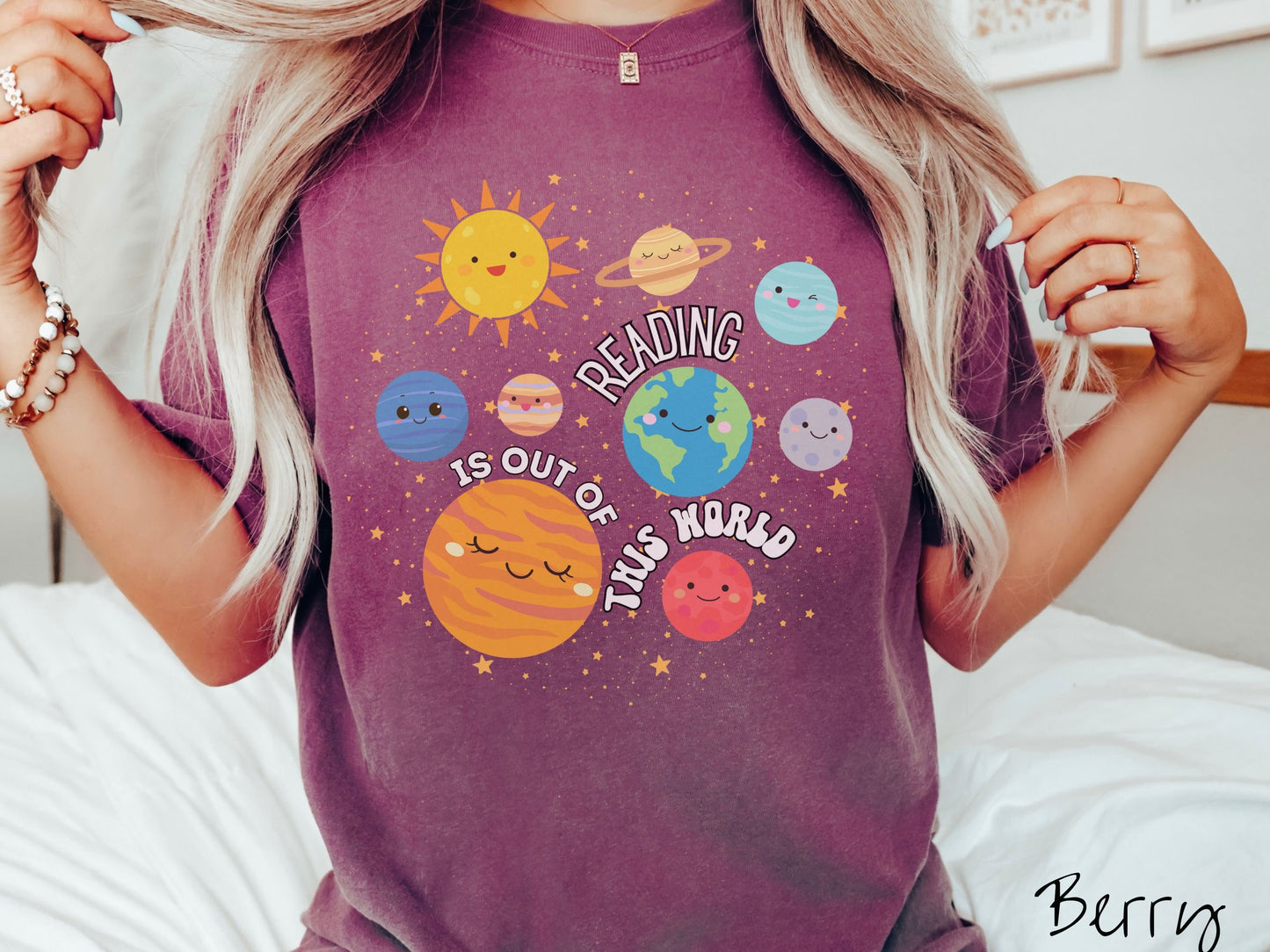 A woman wearing a cute, vintage berry colored shirt showing the solar system and all the planets have smiling, happy faces. There is text saying Reading Is Out of This World among the planets, and there are many stars twinkling in the background.