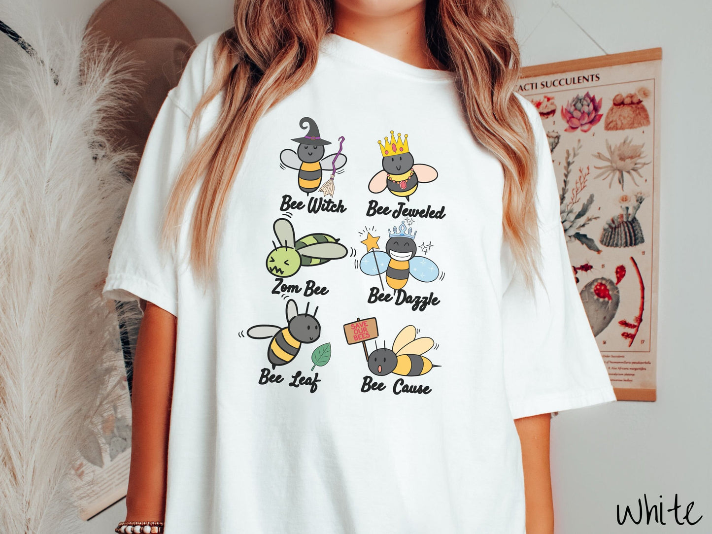 A woman wearing a cute, vintage white colored comfy sweatshirt that has six puns that play on honey bees accompanied with bee drawings. The puns are Bee Witch, Bee Jeweled, Zom Bee, Bee Dazzle, Bee Leaf, and Bee Cause.