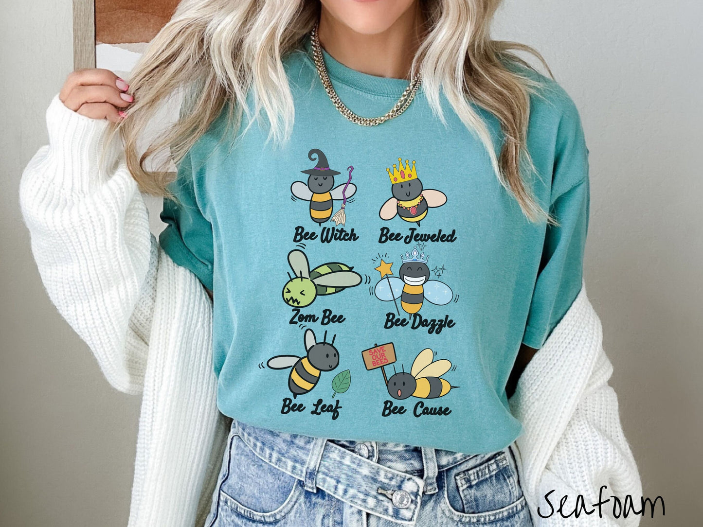 A woman wearing a cute, vintage seafoam colored comfy sweatshirt that has six puns that play on honey bees accompanied with bee drawings. The puns are Bee Witch, Bee Jeweled, Zom Bee, Bee Dazzle, Bee Leaf, and Bee Cause.