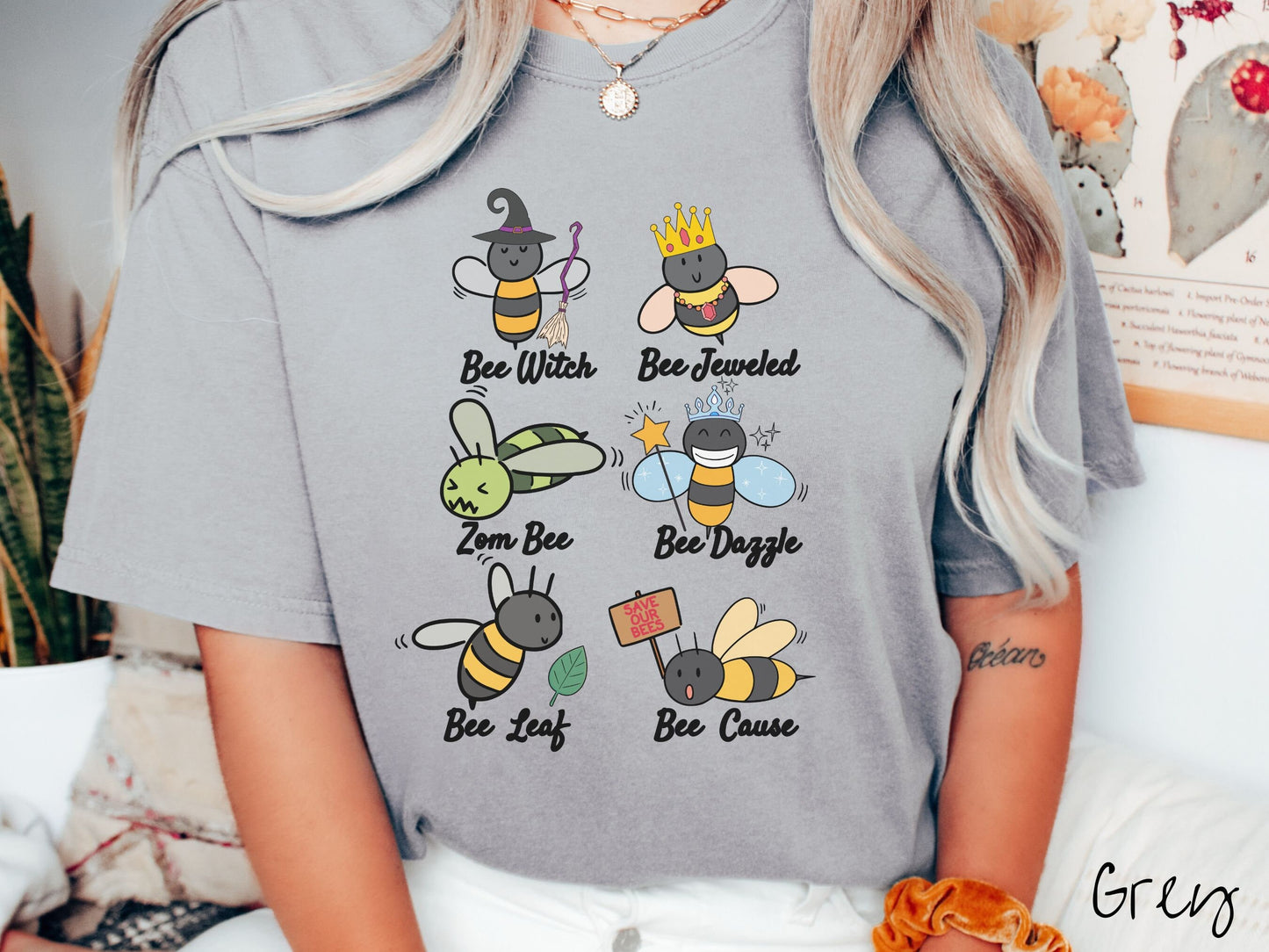 A woman wearing a cute, vintage grey colored comfy sweatshirt that has six puns that play on honey bees accompanied with bee drawings. The puns are Bee Witch, Bee Jeweled, Zom Bee, Bee Dazzle, Bee Leaf, and Bee Cause.
