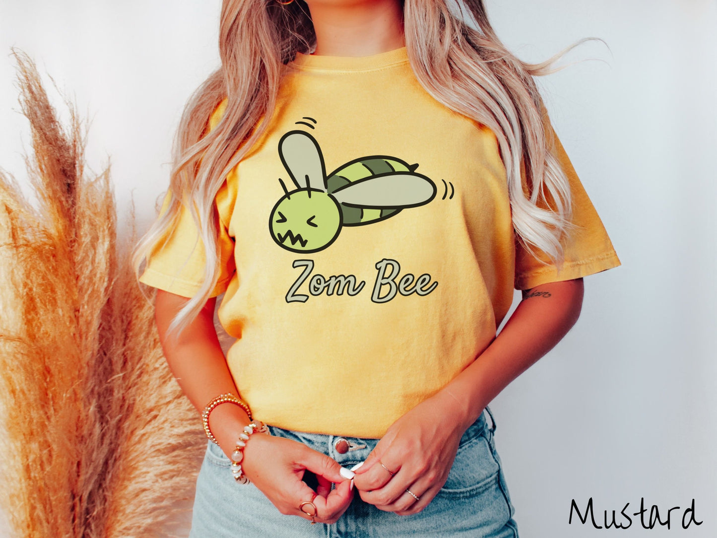A woman wearing a cute, vintage mustard colored Comfort Colors t-shirt with the text Zom Bee in light green, cursive font. Above that is a light and dark green honey bee with a crazy face buzzing in the air.