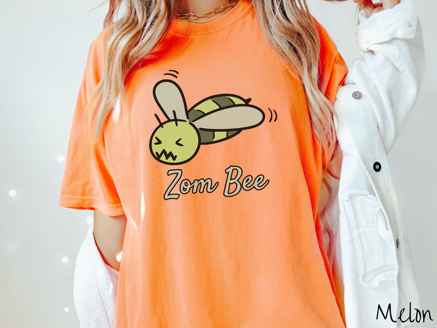 A woman wearing a cute, vintage melon colored Comfort Colors t-shirt with the text Zom Bee in light green, cursive font. Above that is a light and dark green honey bee with a crazy face buzzing in the air.