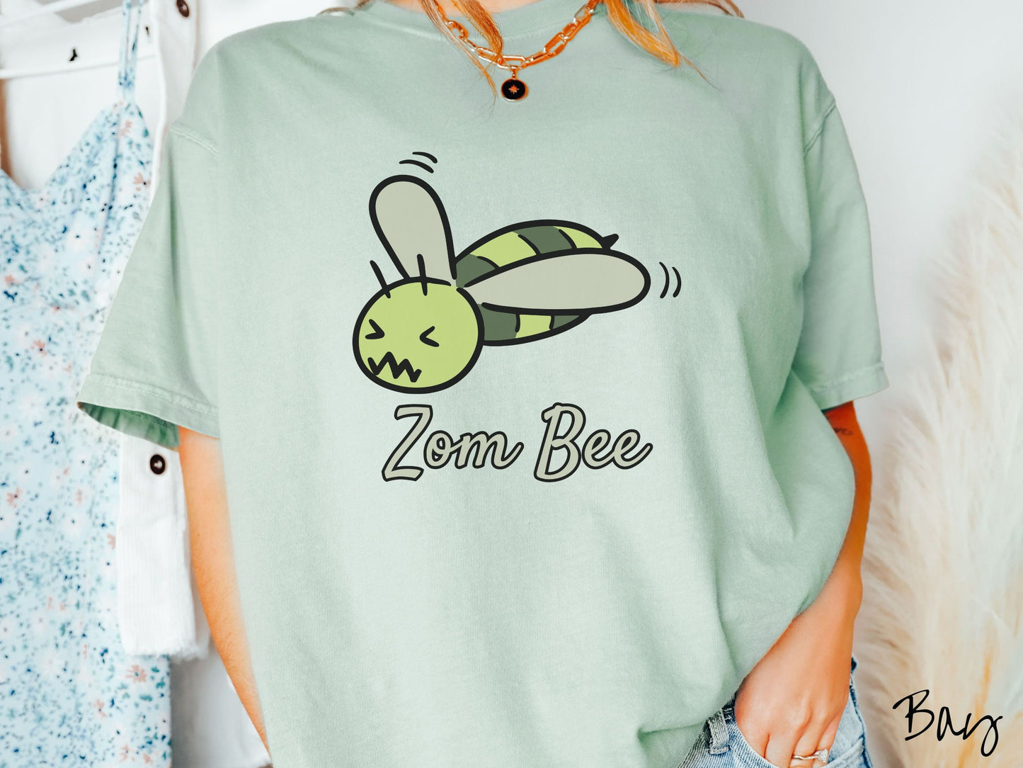A woman wearing a cute, vintage bay colored Comfort Colors t-shirt with the text Zom Bee in light green, cursive font. Above that is a light and dark green honey bee with a crazy face buzzing in the air.