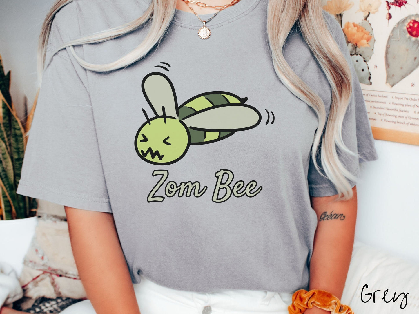A woman wearing a cute, vintage grey colored Comfort Colors t-shirt with the text Zom Bee in light green, cursive font. Above that is a light and dark green honey bee with a crazy face buzzing in the air.