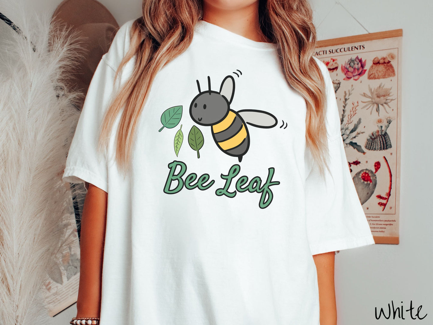 A woman wearing a cute, vintage white colored Comfort Colors t-shirt with the text Bee Leaf in green, cursive font. Above that is a black and yellow honey bee buzzing through the air with green leaves floating next to it.