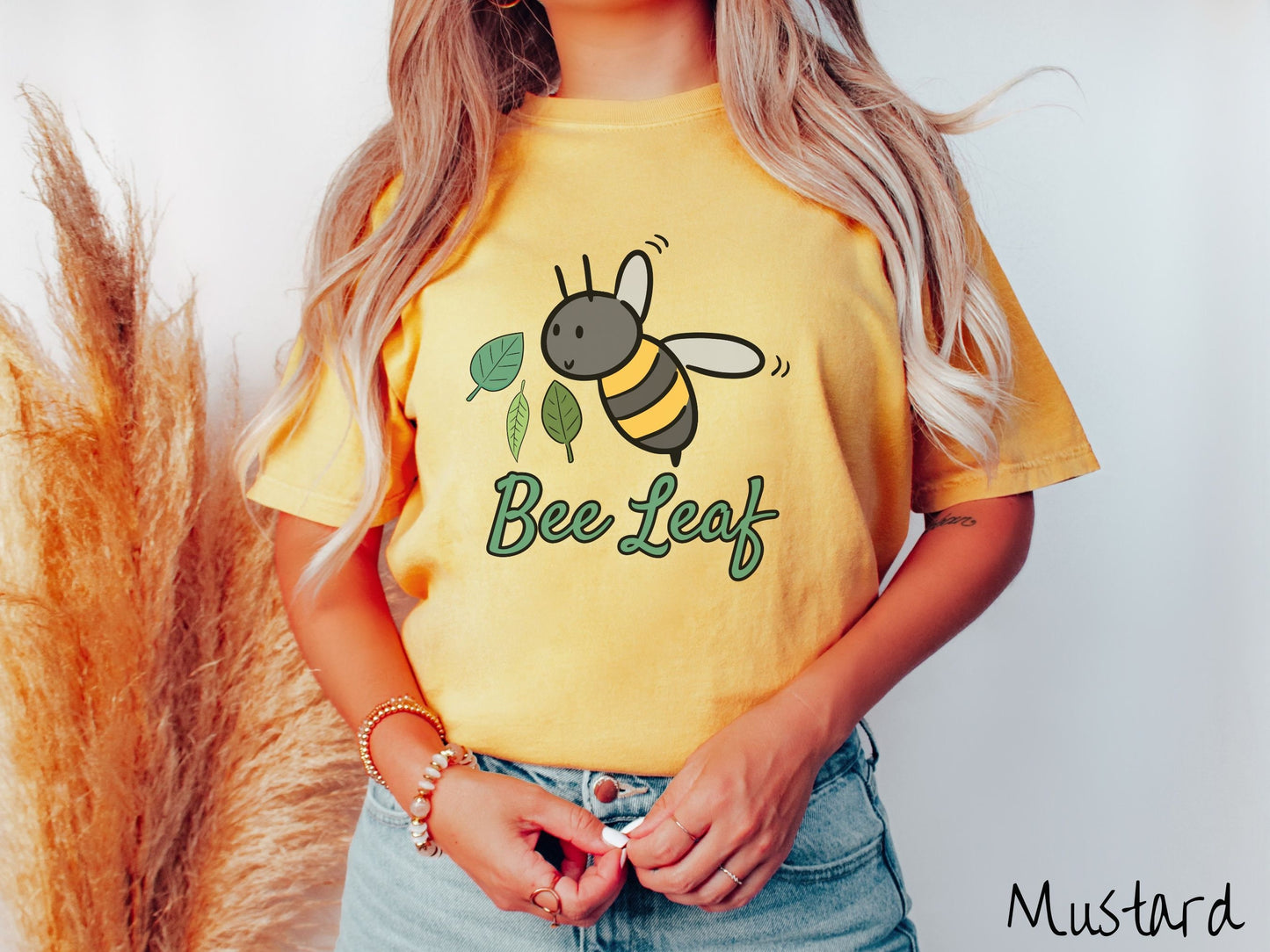 A woman wearing a cute, vintage mustard colored Comfort Colors t-shirt with the text Bee Leaf in green, cursive font. Above that is a black and yellow honey bee buzzing through the air with green leaves floating next to it.