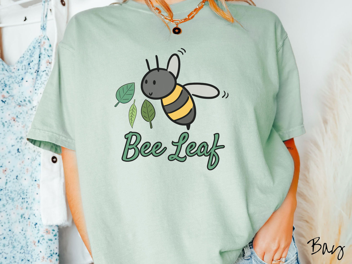 A woman wearing a cute, vintage bay colored Comfort Colors t-shirt with the text Bee Leaf in green, cursive font. Above that is a black and yellow honey bee buzzing through the air with green leaves floating next to it.