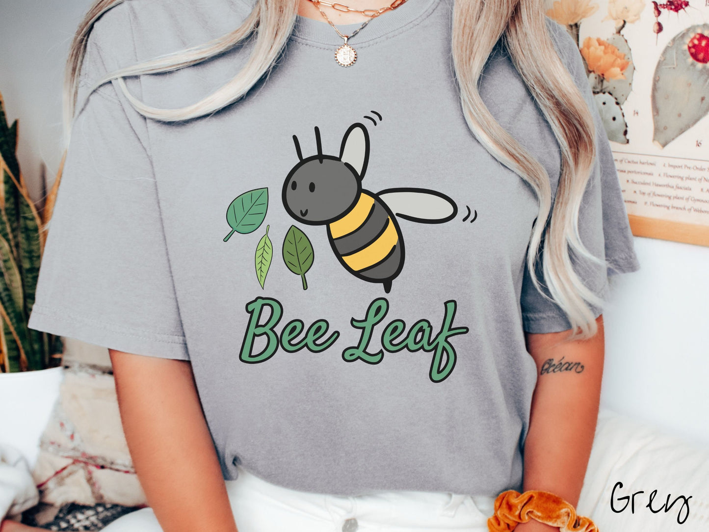 A woman wearing a cute, vintage grey colored Comfort Colors t-shirt with the text Bee Leaf in green, cursive font. Above that is a black and yellow honey bee buzzing through the air with green leaves floating next to it.