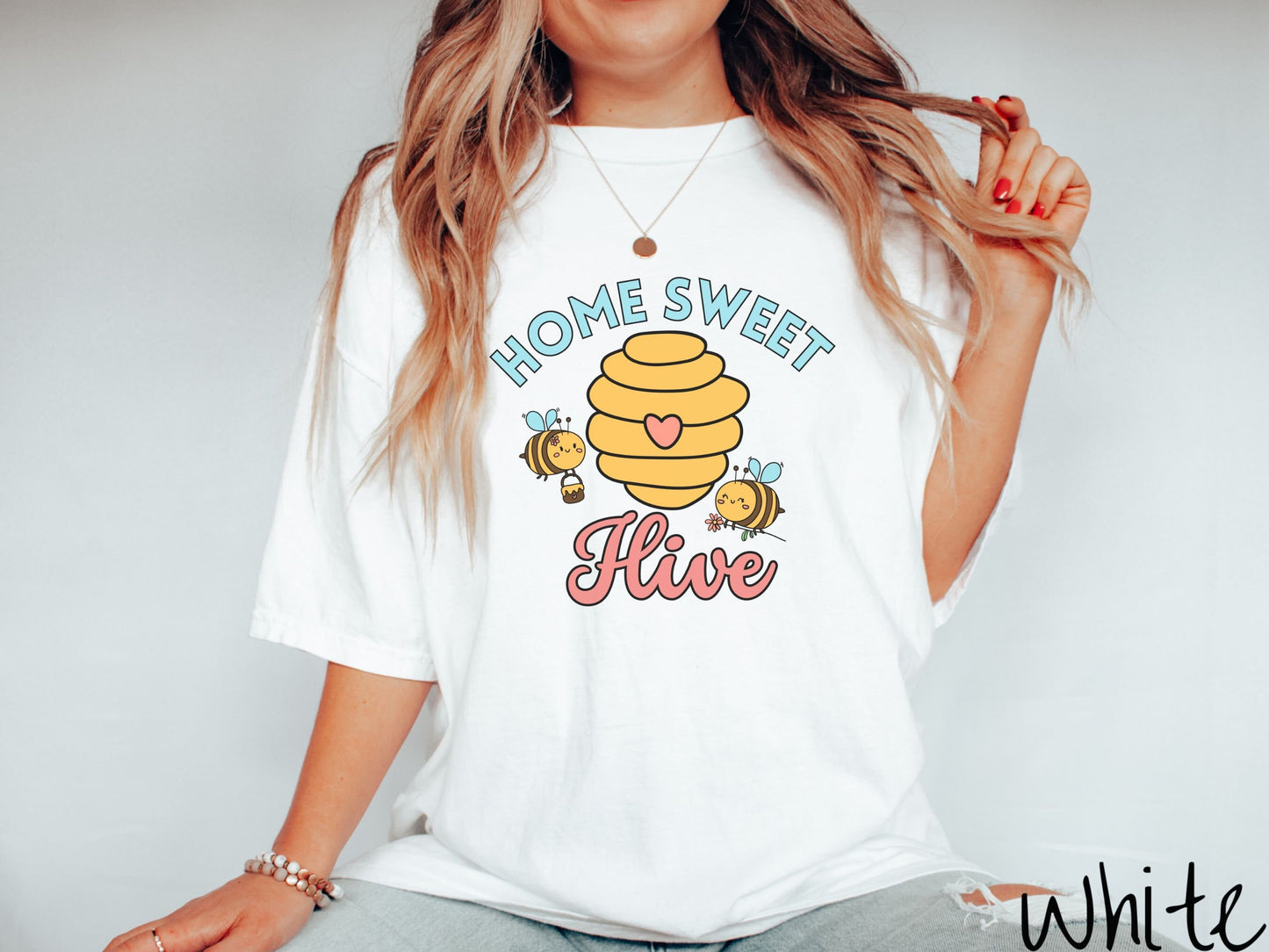 A woman wearing a cute, vintage white colored Comfort Colors t-shirt with the text Home Sweet Hive in cyan and red font. In between the words are a yellow bee hive with a red heart on it and smiling honey bees flying around holding flowers.