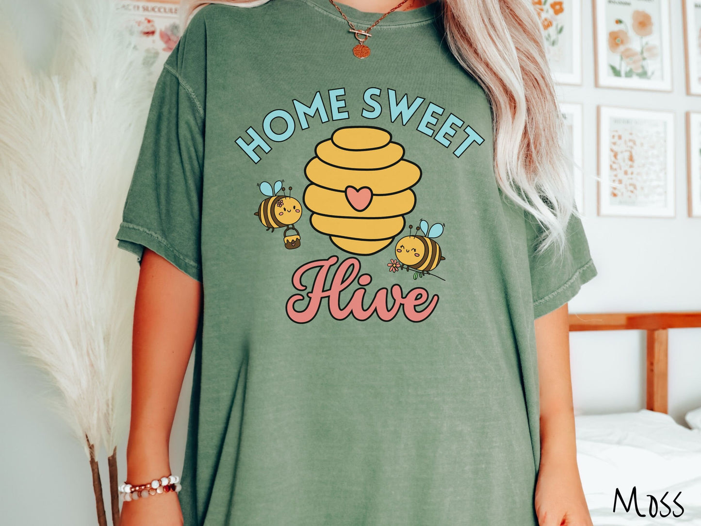 A woman wearing a cute, vintage moss colored Comfort Colors t-shirt with the text Home Sweet Hive in cyan and red font. In between the words are a yellow bee hive with a red heart on it and smiling honey bees flying around holding flowers.