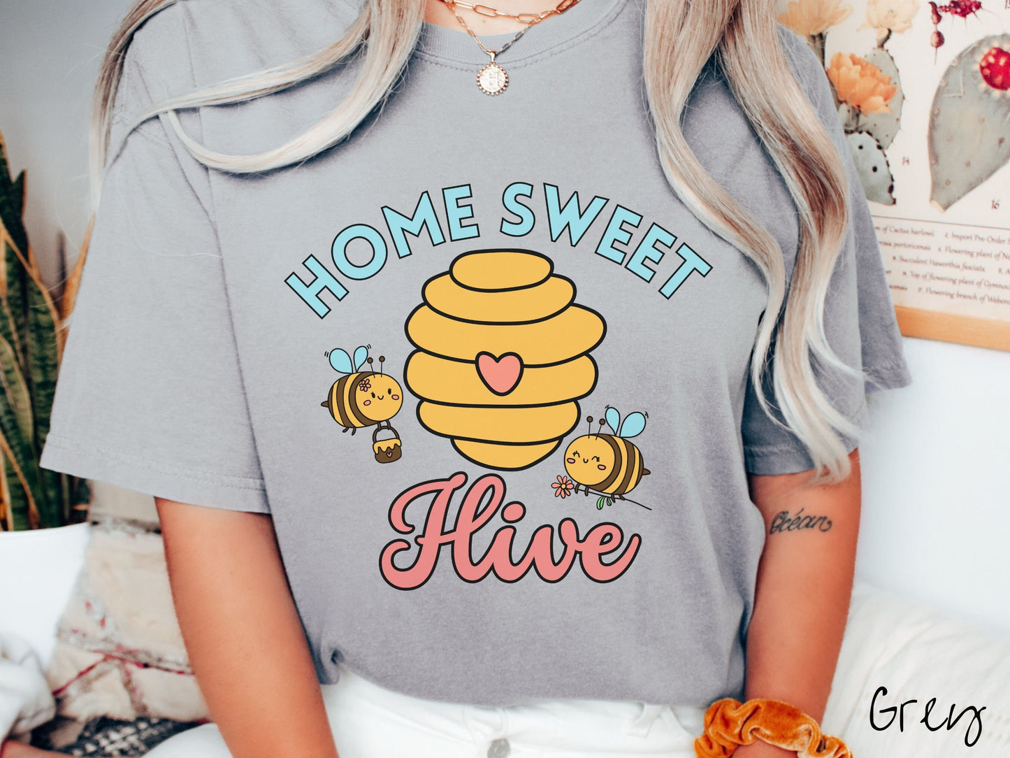 A woman wearing a cute, vintage grey colored Comfort Colors t-shirt with the text Home Sweet Hive in cyan and red font. In between the words are a yellow bee hive with a red heart on it and smiling honey bees flying around holding flowers.