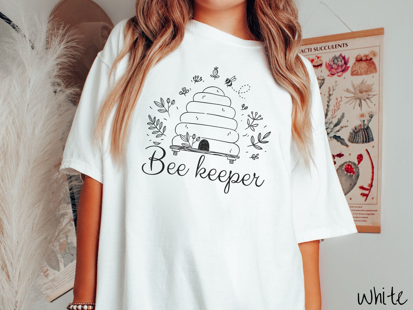 A woman wearing a cute, vintage white colored Comfort Colors t-shirt with the text Beekeeper across the front in black, cursive font. Above this is a black-outlined bee hive with bees buzzing around it and there are leaves in the air.