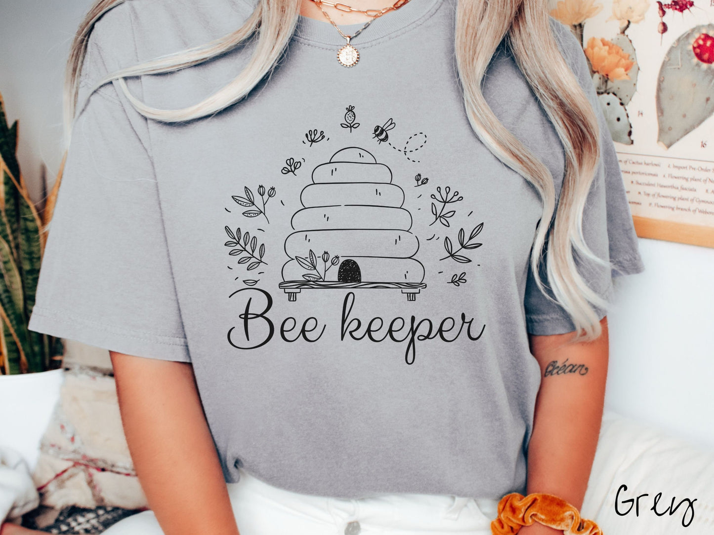 A woman wearing a cute, vintage grey colored Comfort Colors t-shirt with the text Beekeeper across the front in black, cursive font. Above this is a black-outlined bee hive with bees buzzing around it and there are leaves in the air.