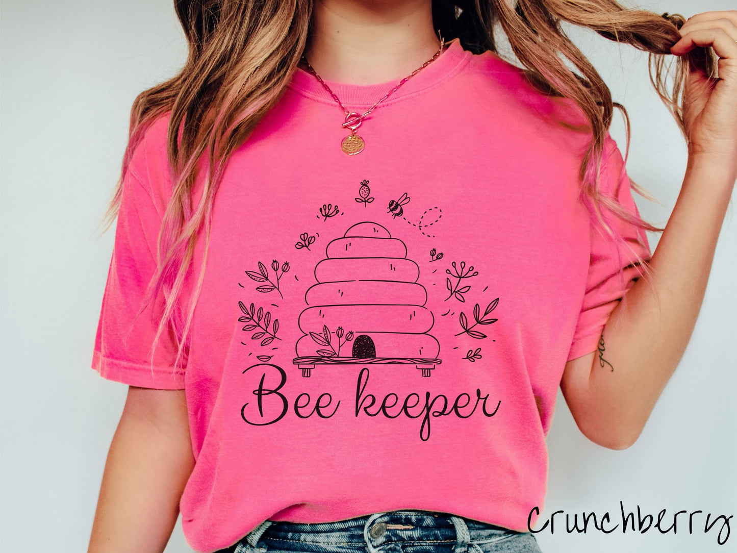 A woman wearing a cute, vintage crunchbery colored Comfort Colors t-shirt with the text Beekeeper across the front in black, cursive font. Above this is a black-outlined bee hive with bees buzzing around it and there are leaves in the air.