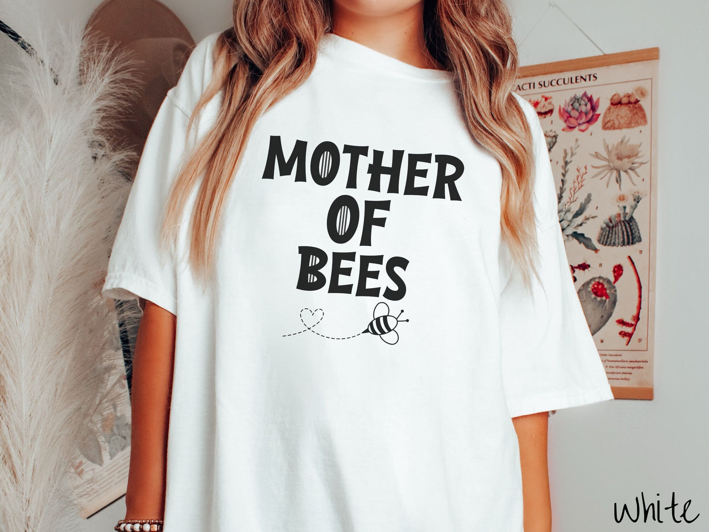 A woman wearing a cute, vintage white colored Comfort Colors t-shirt with the text Mother of Bees in black, uppercase font. Below that is a black outlined bee buzzing around, the trail it leaves has created a heart shape in the air.