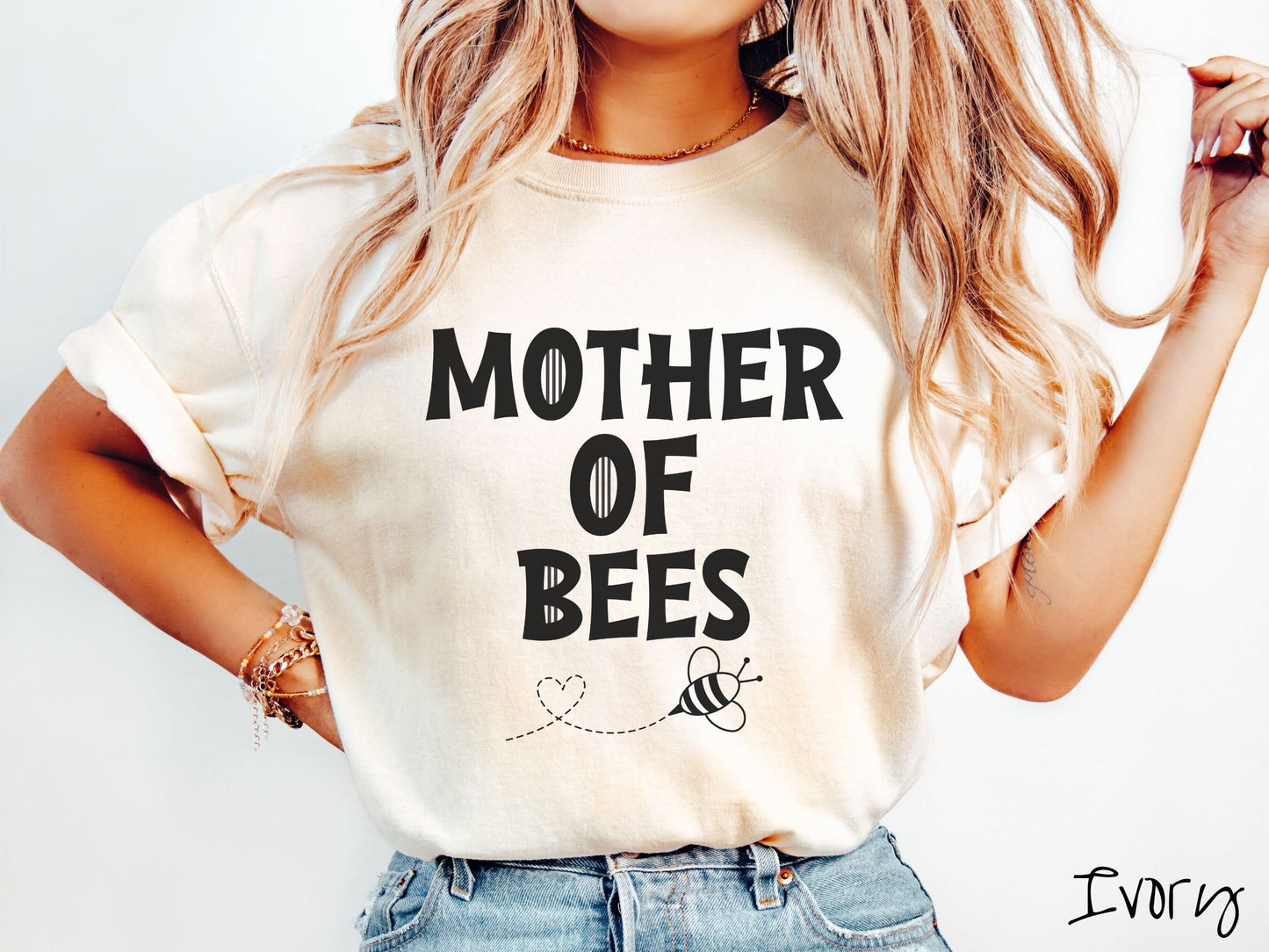 A woman wearing a cute, vintage ivory colored Comfort Colors t-shirt with the text Mother of Bees in black, uppercase font. Below that is a black outlined bee buzzing around, the trail it leaves has created a heart shape in the air.
