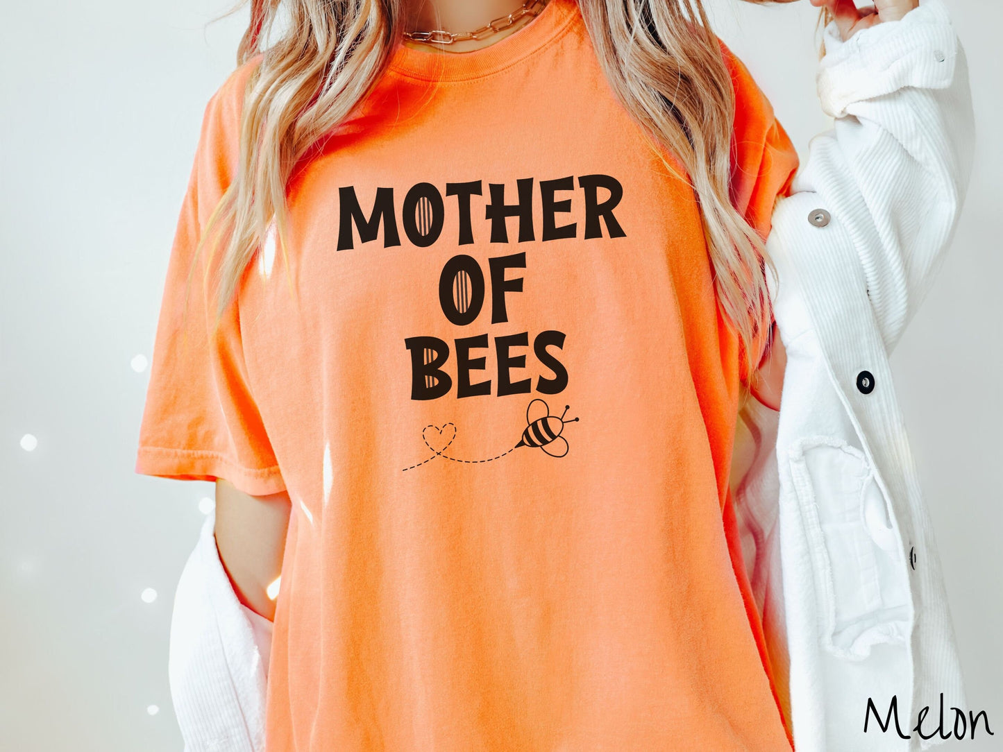 A woman wearing a cute, vintage melon colored Comfort Colors t-shirt with the text Mother of Bees in black, uppercase font. Below that is a black outlined bee buzzing around, the trail it leaves has created a heart shape in the air.