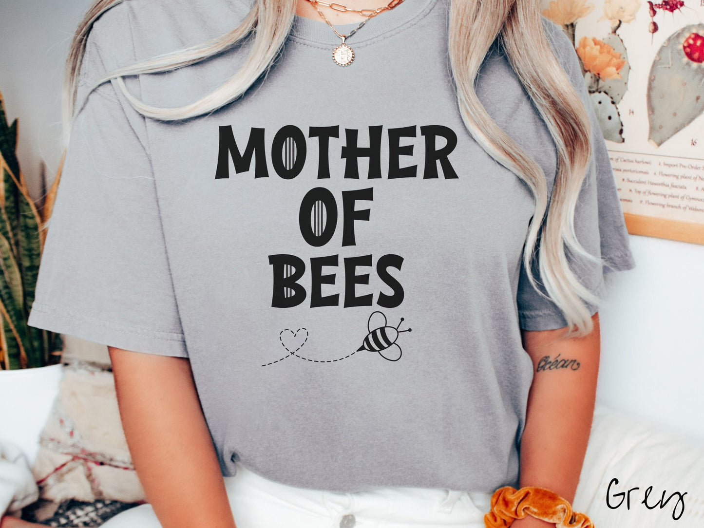 A woman wearing a cute, vintage grey colored Comfort Colors t-shirt with the text Mother of Bees in black, uppercase font. Below that is a black outlined bee buzzing around, the trail it leaves has created a heart shape in the air.