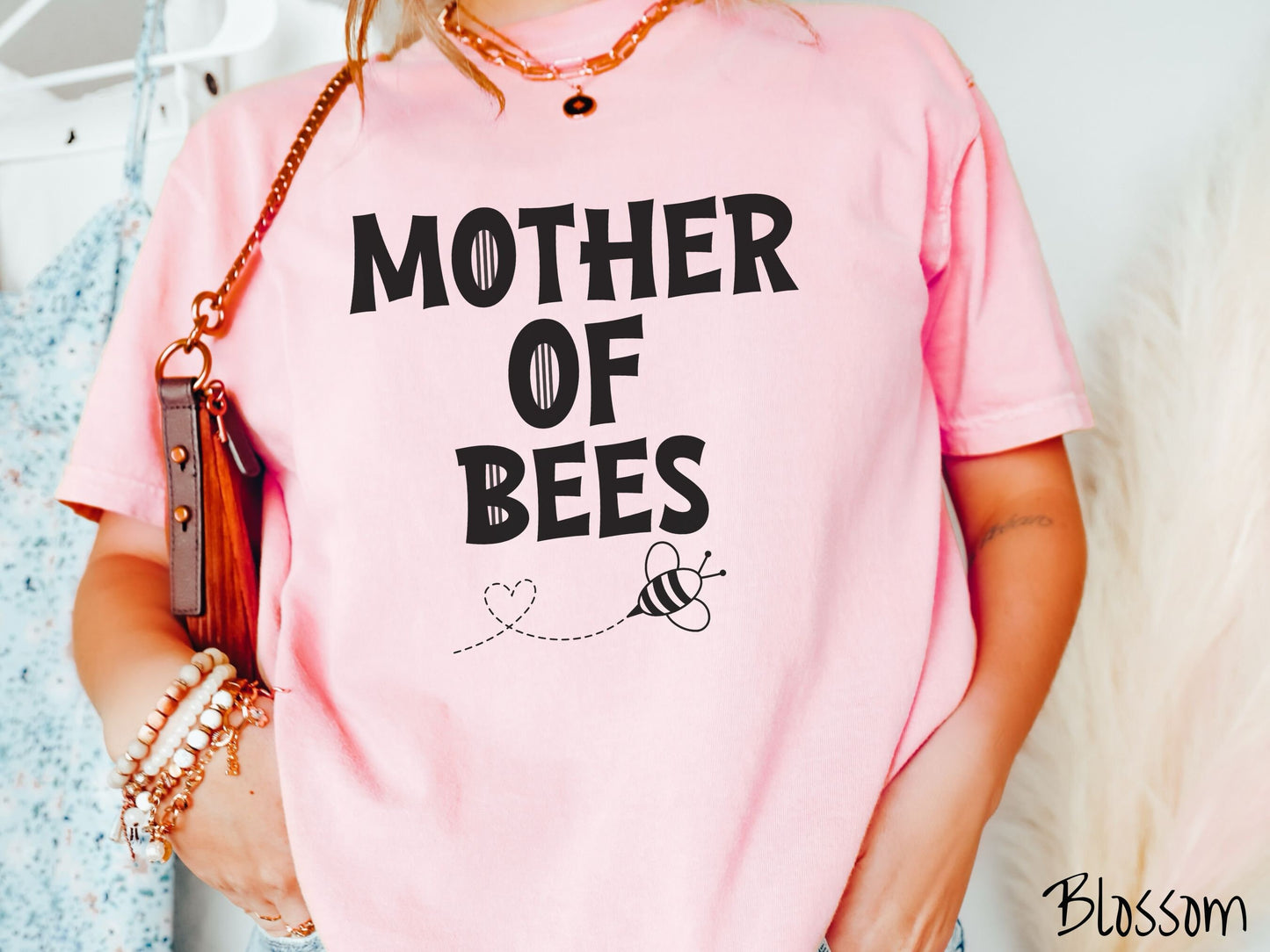 A woman wearing a cute, vintage blossom colored Comfort Colors t-shirt with the text Mother of Bees in black, uppercase font. Below that is a black outlined bee buzzing around, the trail it leaves has created a heart shape in the air.