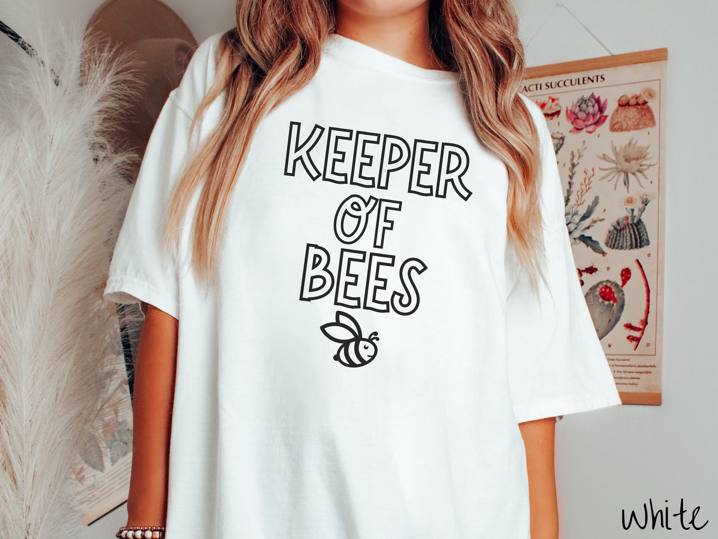 A woman wearing a cute, vintage white colored Comfort Colors t-shirt with the text Keeper of Bees in black uppercase font. Below the text is a black-outlined smiling bee looking up.