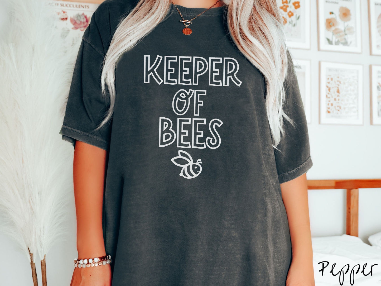 A woman wearing a cute, vintage pepper colored Comfort Colors t-shirt with the text Keeper of Bees in white uppercase font. Below the text is a white-outlined smiling bee looking up.