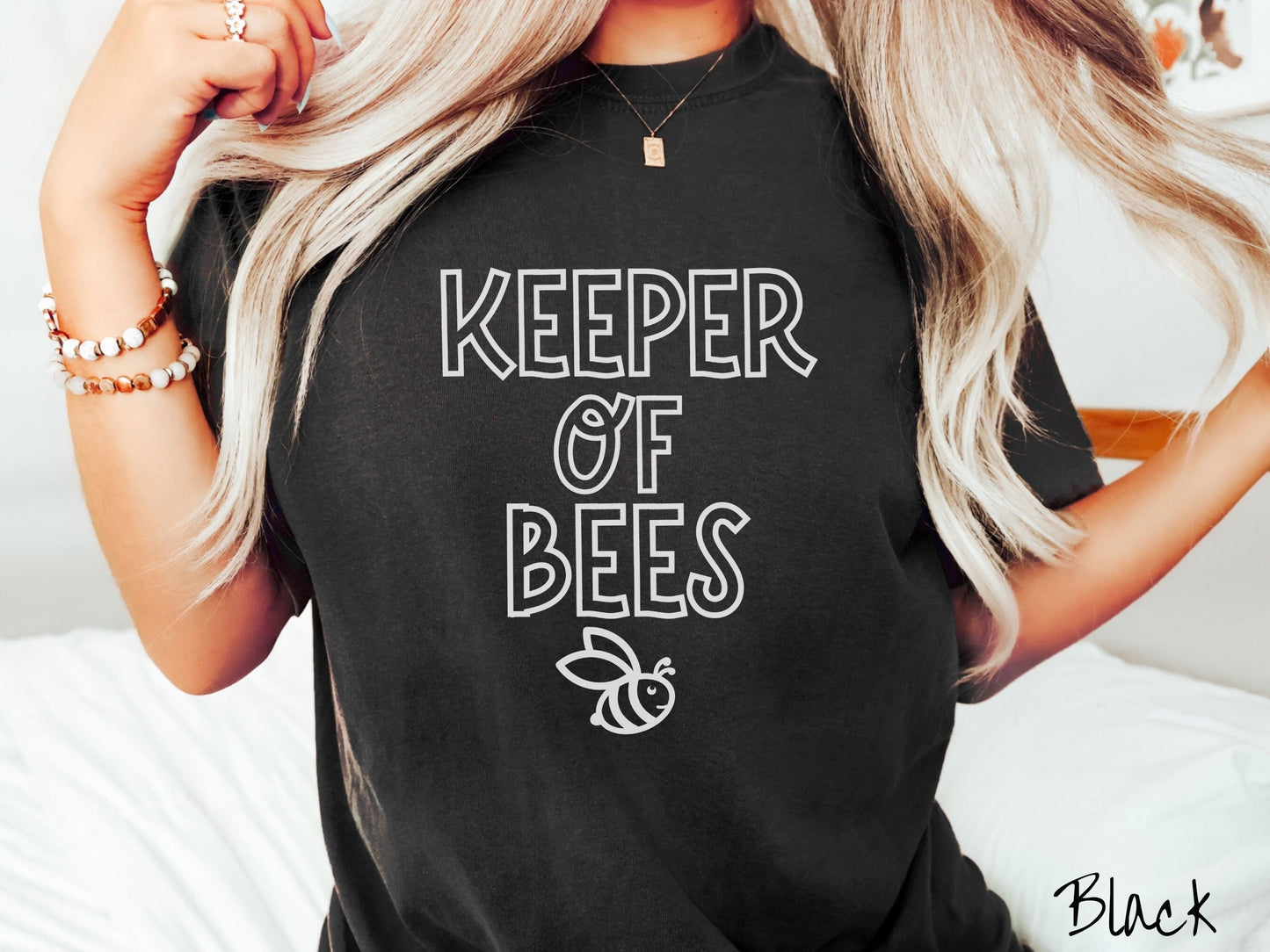 A woman wearing a cute, vintage black colored Comfort Colors t-shirt with the text Keeper of Bees in white uppercase font. Below the text is a white-outlined smiling bee looking up.