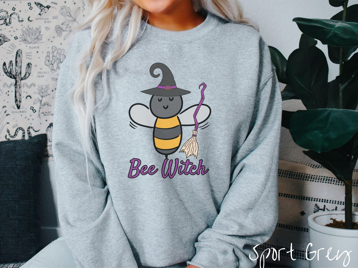 A woman wearing a cute, vintage sport grey colored comfy sweatshirt with the text Bee Witch in purple, cursive font. Above that is a black and yellow honey bee holding a purple, straw broom and wearing a black witch hat with a purple hat band.