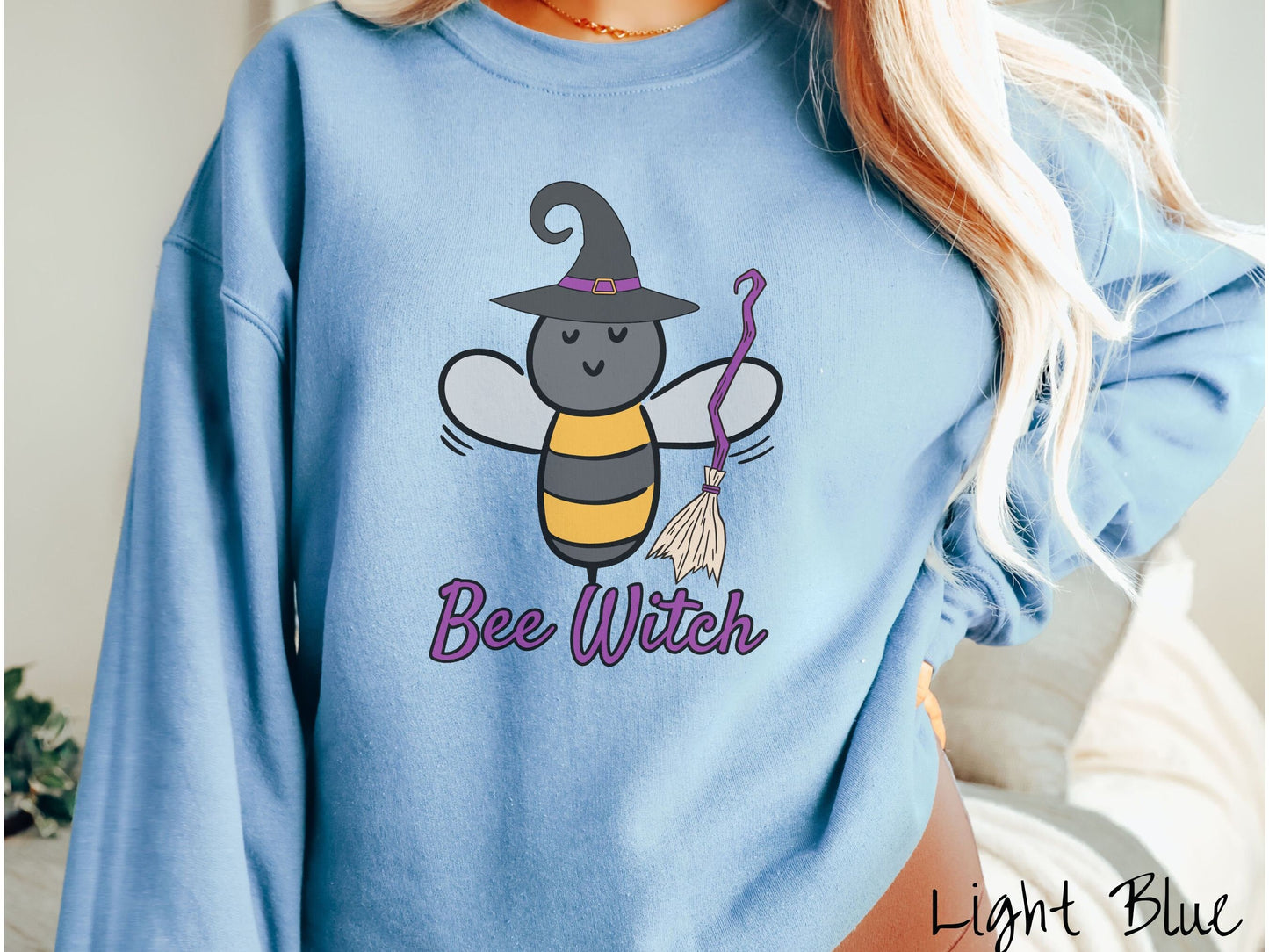 A woman wearing a cute, vintage light blue colored comfy sweatshirt with the text Bee Witch in purple, cursive font. Above that is a black and yellow honey bee holding a purple, straw broom and wearing a black witch hat with a purple hat band.