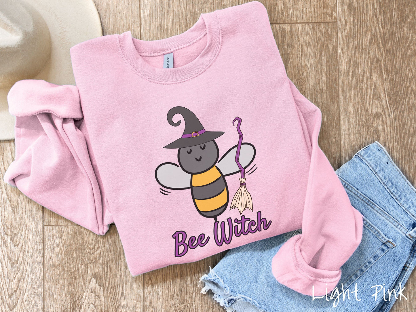 A cute, vintage light pink colored comfy sweatshirt with the text Bee Witch in purple, cursive font. Above that is a black and yellow honey bee holding a purple, straw broom and wearing a black witch hat with a purple hat band.
