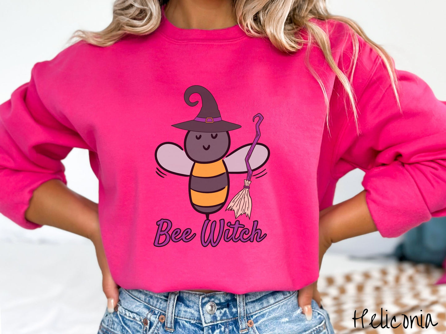 A woman wearing a cute, vintage heliconia colored comfy sweatshirt with the text Bee Witch in purple, cursive font. Above that is a black and yellow honey bee holding a purple, straw broom and wearing a black witch hat with a purple hat band.