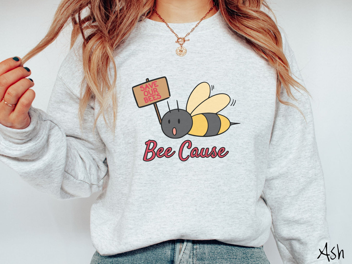 A woman wearing a cute, vintage ash colored comfy sweatshirt with the text Bee Cause in red, cursive font. Above that is a black and yellow honey bee speaking with its mouth open and holding a sign that reads Save Our Bees in red font.